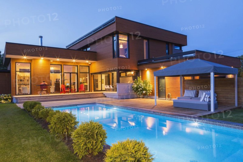 Rear of modern cube style home with stained horizontal wood cladding, in-ground swimming pool and wooden deck with gazebo canopy at dusk, Quebec, Canada, North America