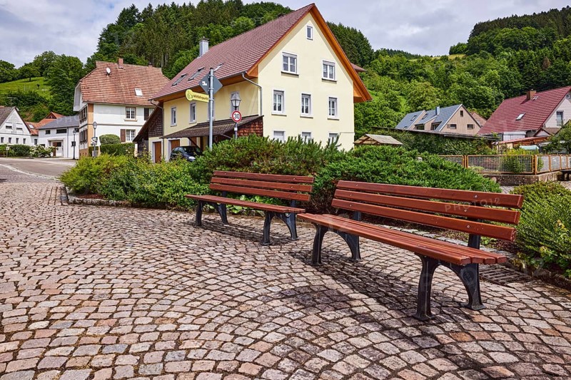 Benches, pavement made of cobblestones, car park, multi-family houses, hilly landscape with coniferous forest, overcast sky with Nimbostratus clouds, sunlight, frog perspective, Bollenbacher Straße, Haslach im Kinzigtal, Black Forest, Ortenaukreis, Baden-Württemberg, Germany, Europe