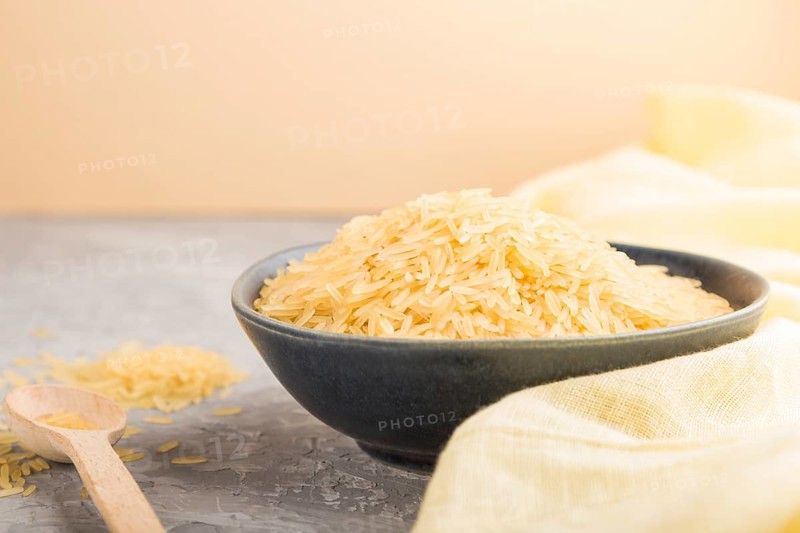 Blue ceramic bowl with raw golden rice and wooden spoon on a gray concrete background and yellow textile. Side view, close up, selective focus