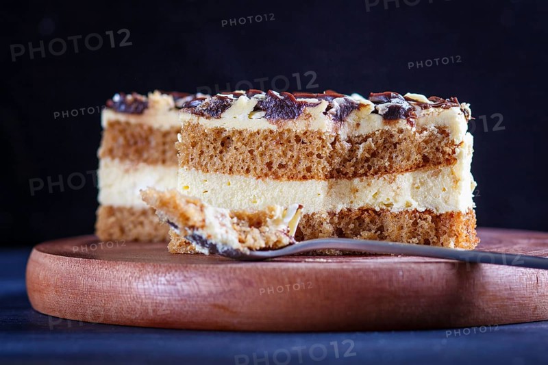 A piece of cake with milk and butter cream cutted with spoon on a wooden kitchen board, close up, selective focus, black background