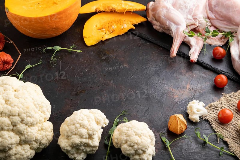 Whole raw rabbit with pumpkin, tomatoes, cauliflower pea sprouts on a black concrete background and linen textile. Side view, copy space