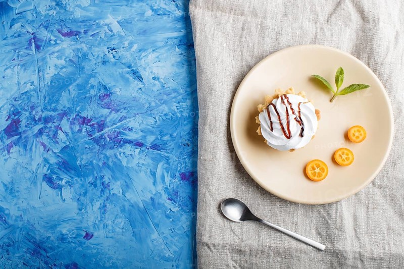 Cake with whipped egg cream on a light brown plate with kumquat slices and mint leaves on a blue concrete background with linen napkin. copy space, flat lay, top view
