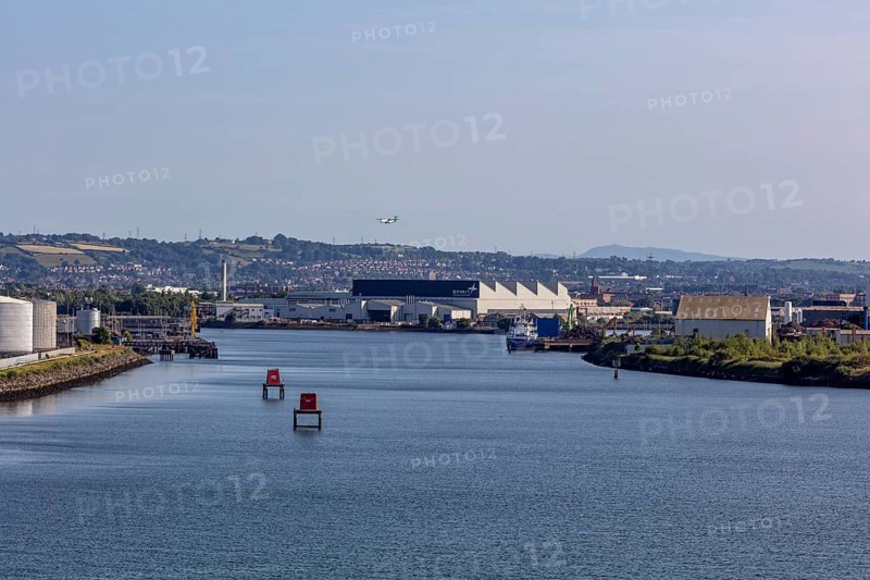 River landscape with industrial buildings, buoys and a flying aeroplane under a blue sky, Belfast