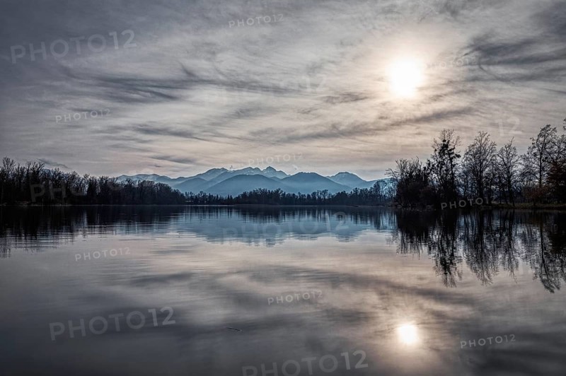 Still lake with mountain reflection in the water, surrounded by bare trees and overcast sky with sunspots, Happinger See