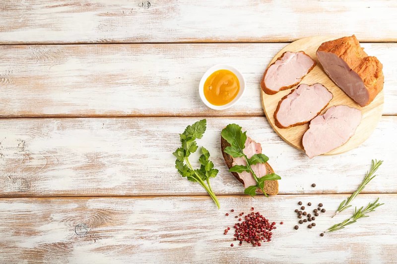 Smoked pork ham on cutting board with pepper and herbs on white wooden background. Top view, flat lay, copy space