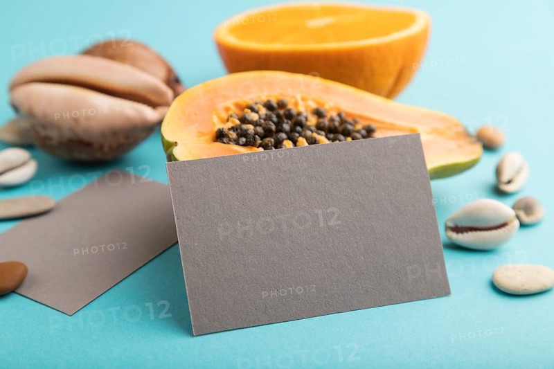 Gray paper business card with ripe cut papaya, orange, seashells on blue pastel pastel background. Side view, copy space. Tropical, healthy food, vacation, holidays concept