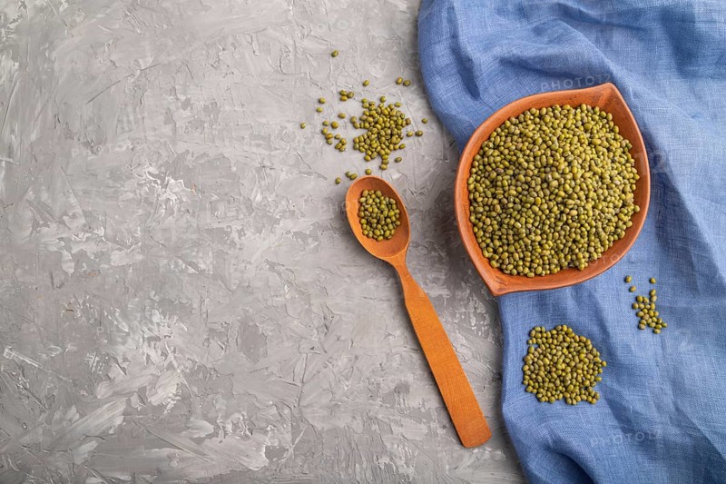 Clay bowl with raw green mung bean and wooden spoon on a gray concrete background and blue textile. Top view, flat lay, copy space