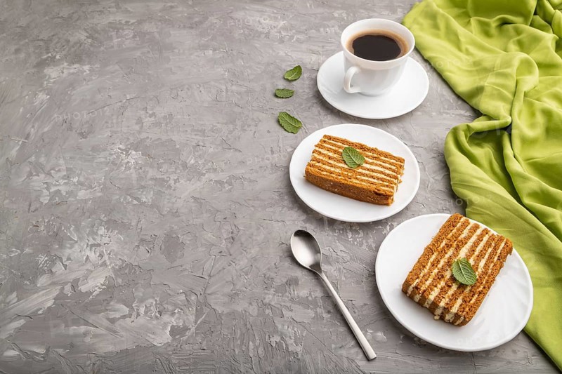 Homemade honey cake with milk cream and mint with cup of coffee on a gray concrete background and green textile. Side view, copy space