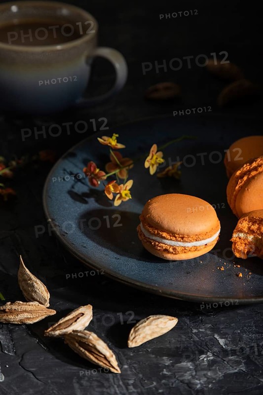Orange macarons or macaroons cakes with cup of coffee on a black concrete background. Hard light, contrast, low key. Side view, close up