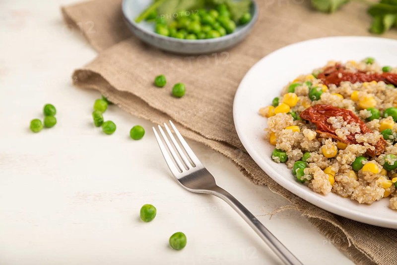 Quinoa porridge with green pea, corn and dried tomatoes on ceramic plate on a white wooden background and linen textile. Side view, close up, selective focus