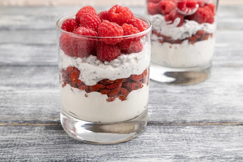 Yogurt with raspberry, goji berries and chia seeds in glass on gray wooden background. Side view, close up, selective focus