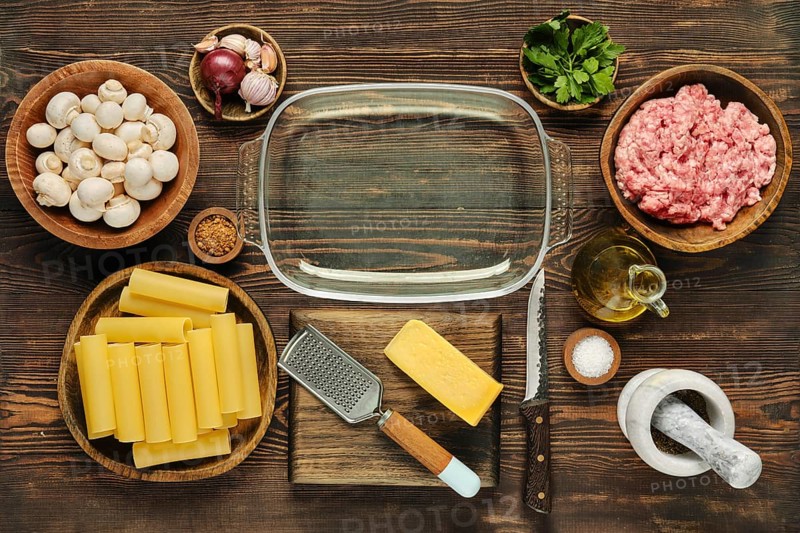 Fresh ingredients are arranged neatly on a wooden countertop in preparation for a pasta meal. Components include mushrooms, ground meat, pasta sheets, cheese, garlic, and herbs