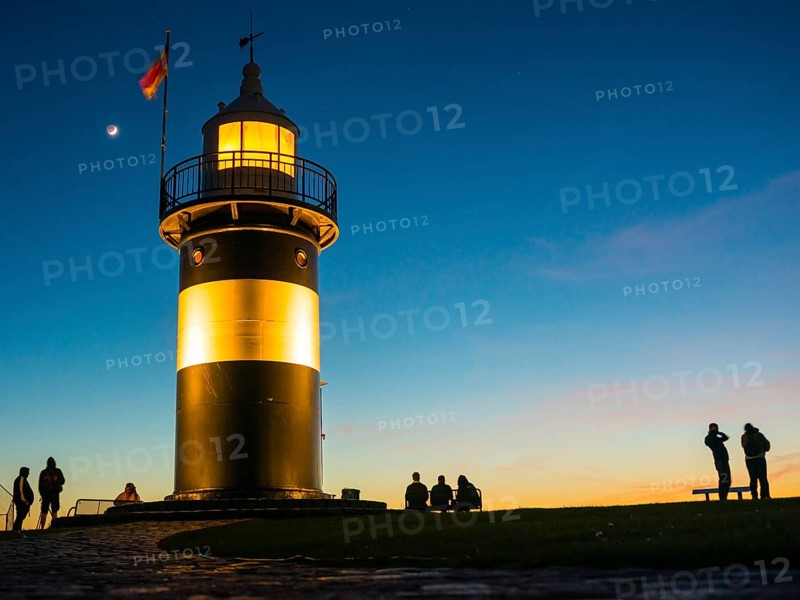 Black and white lighthouse, called 'Kleiner Preuße', illuminated at dusk with a clear blue sky, a rising moon and silhouettes of people standing and sitting next to it, maritime, German flag waving in the wind, bright beacon, paved curved path, twilight, evening, dusk, maritime, calm atmosphere, North Sea resort Wremen, Lower Saxony Wadden Sea National Park, World Heritage Site, Wurster North Sea coast, Land Wursten, district of Cuxhaven, Lower Saxony, Germany, Europe
