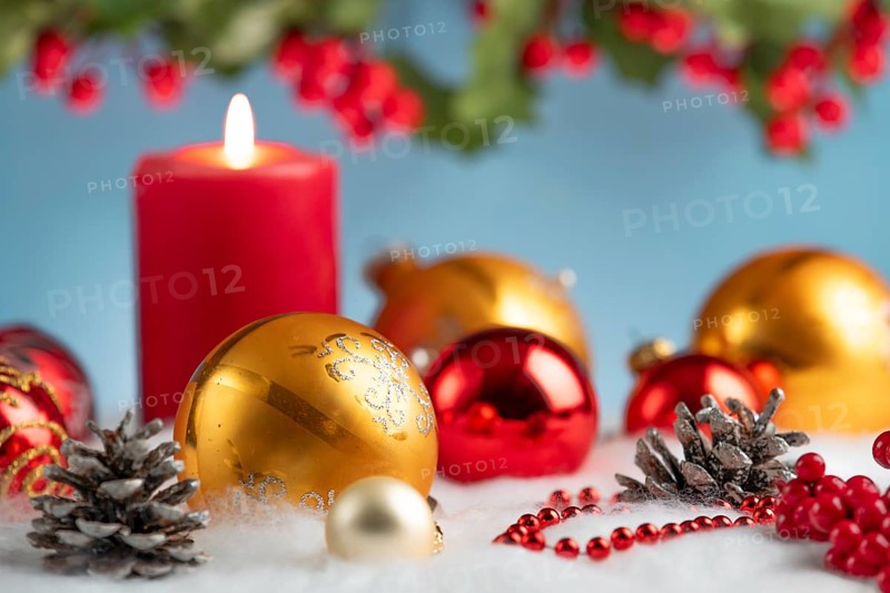 Red and gold Christmas baubles next to a lit candle on artificial snow, decorative festive atmosphere