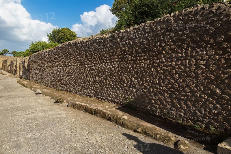 Long compacted crushed stone paved street with sidewalk and old stone walls plus residential building structures at the ancient ruins of Pompeii, Campania region, Italy, Europe