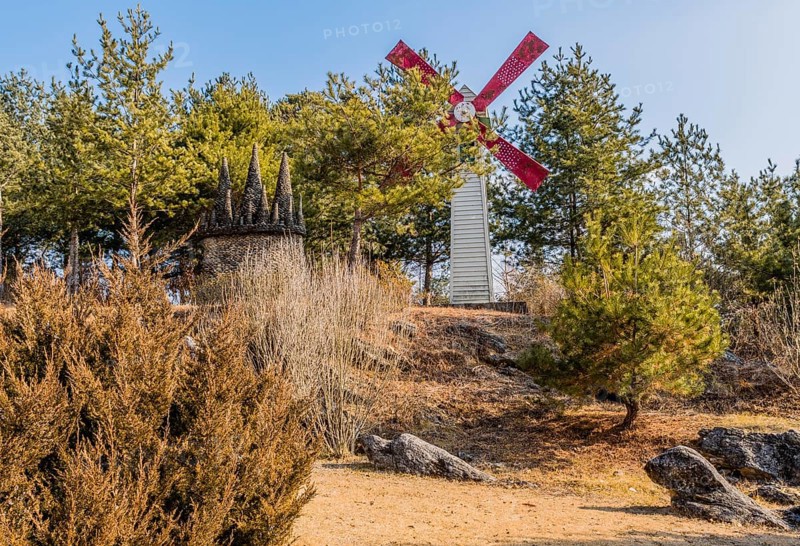 Gangwon-Do, South Korea. January 12, 2020: Windmill next to miniature rock castle surrounded by evergreen trees located in public park on sunny afternoon