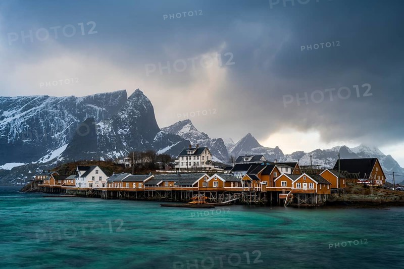 Traditional yellow rorbuer cabins on Sakrisøy Island, snow-capped rocky mountains in the back, Sakrisoy, Reine, Lofoten, Norway, Europe