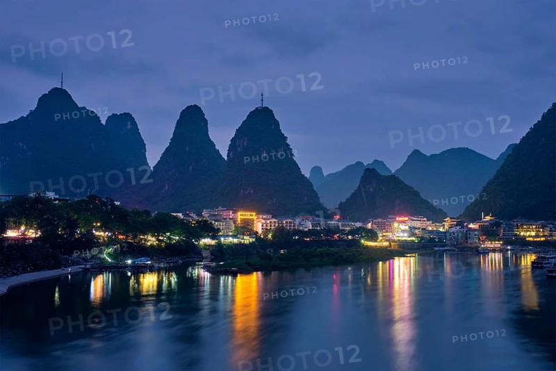 View of Yangshuo town illuminated in the evening with dramatic karst mountain landscape in background over Li river. Yangshuo, China, Asia