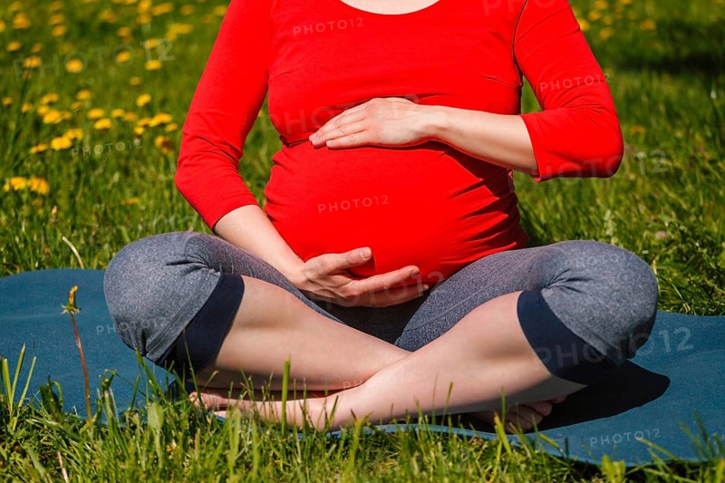 Pregnancy yoga exercise, pregnant woman doing asana Sukhasana easy yoga pose holding her abdomen outdoors on grass lawn with dandelions in summer