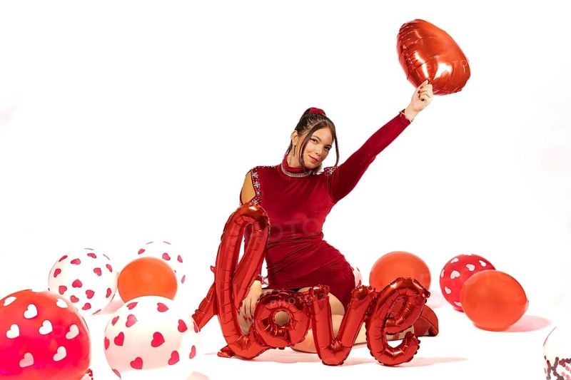Beautiful elegant girl in red dress surrounded by balloons on white background with love sign. Valentine's day concept. Red and white color palette