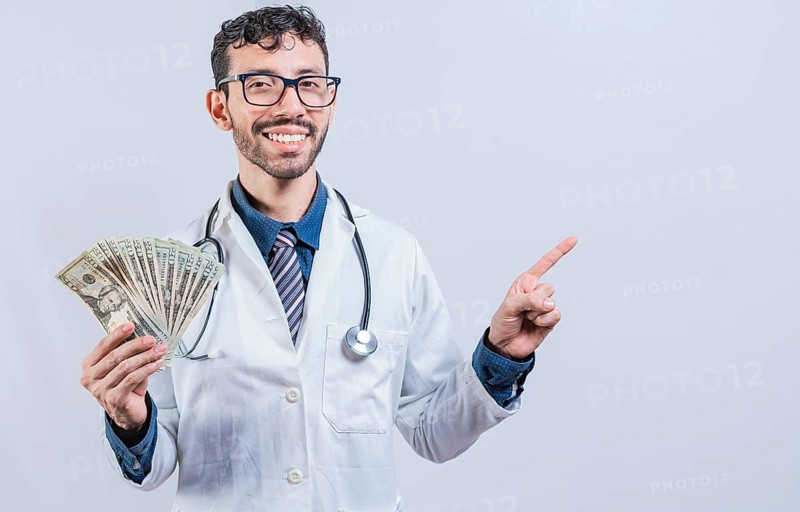 Happy young doctor holding money and pointing to side. Handsome smiling doctor showing money and pointing side. Latin doctor