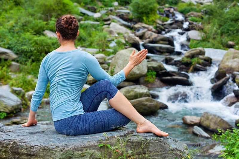 Yoga exercise outdoors, woman doing Ardha matsyendrasanaasana asana, half spinal twist pose at tropical waterfall in Himalayas in India