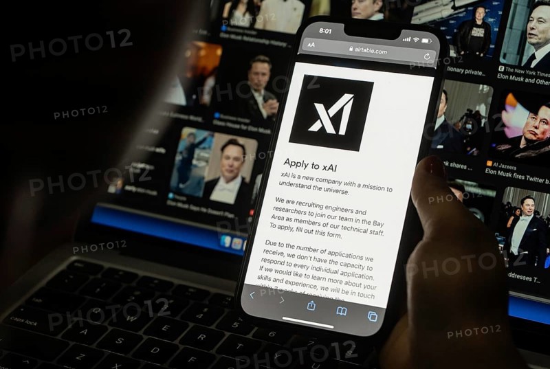 In this photo illustration, A person browses website of xAI, displayed on a mobile phone, Elon Musk images in the background screen, in Guwahati, India 0n 15 July 2023. Tesla CEO Elon Musk launches Artificial Intelligence company xAI on 12 July 2023, the company's goal is to understand the true nature of the universe