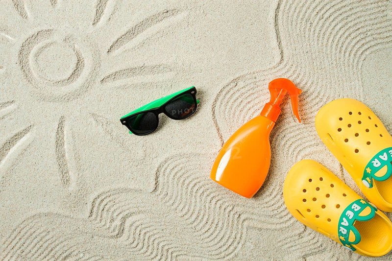Drawing on the sand, sun and waves, sunglasses, with an orange bottle, yellow rubber slippers, sun spray, on the sand, top view, no people