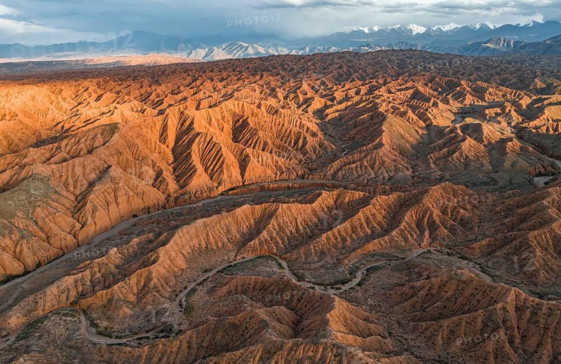 River bed runs through a landscape of eroded hills, badlands at sunset, mountain peaks of the Tian Shan Mountains in the background, aerial view, Canyon of the Forgotten Rivers, Issyk Kul, Kyrgyzstan, Asia