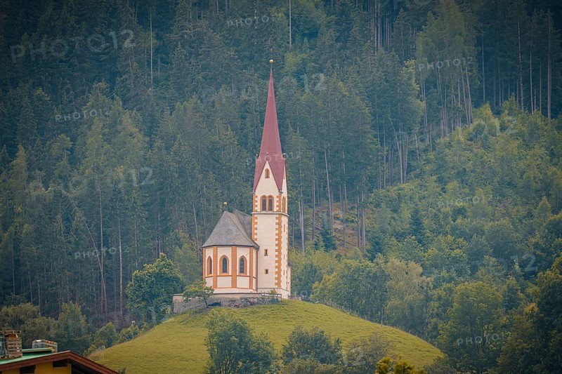 A church with a high tower stands on a green hill in front of a dense forest, Mayrhofen, Zillertal, Austria, Europe