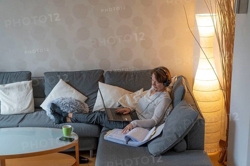 Woman, mid-50s, works from home, with laptop and communicates with colleagues via headset, home office, on the sofa