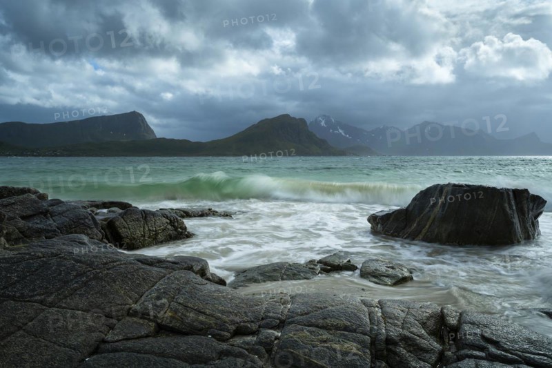 Seascape on the beach at Haukland. View of the mountains of Vestvagoya and Myrland on Flakstadoya. Offersoykammen on the left. Clouds in the sky, some sun. Rocks in the foreground. A wave in the sea. Early summer. Long exposure. Haukland Beach, Haukland, Vestvagoya, Lofoten, Norway, Europe