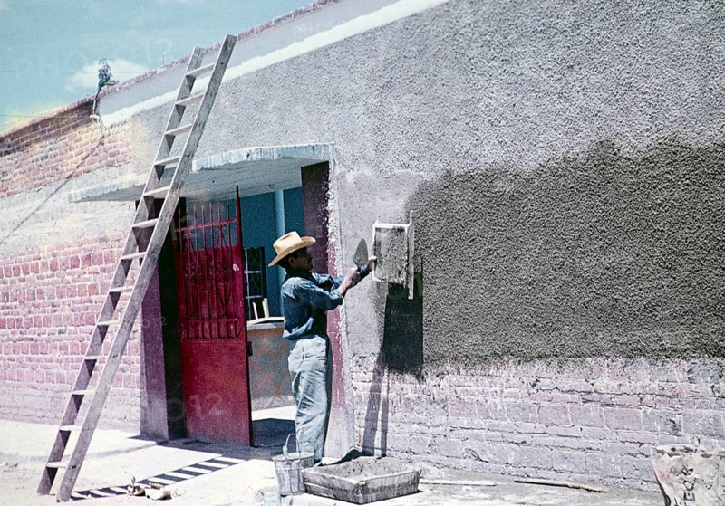 Construction worker throwing cement through a sieve to make wall panelling over modern red bricks, Mexico around 1961