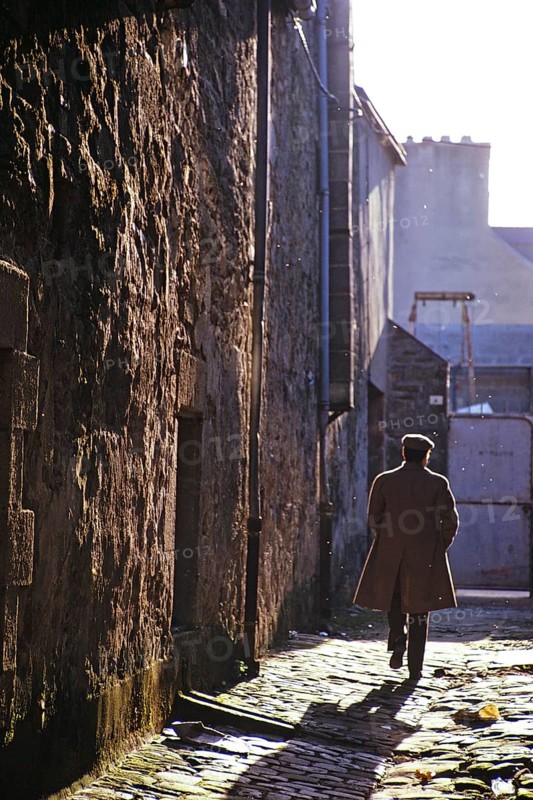 Rückansicht eines Mannes mit Mantel und flacher Mütze, der eine gepflasterte Gasse in Quimper, Bretagne, Frankreich, 1974, entlanggeht