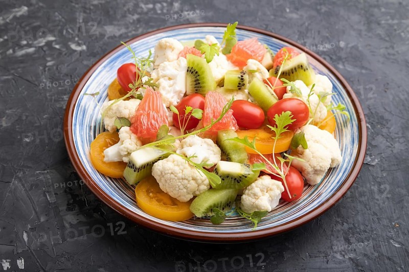 Vegetarian salad of cauliflower cabbage, kiwi, tomatoes, microgreen sprouts on black concrete background. Side view, close up