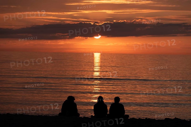 Three people sitting on the beach watching the sunset over the sea, silhouettes in front of warm light, Baltic Sea peninsula Fischland-Darß-Zingst, Dierhagen, Mecklenburg-Vorpommern, Germany, Europe
