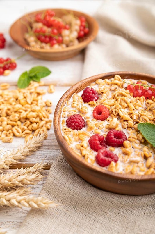 Wheat flakes porridge with milk, raspberry and currant in wooden bowl on white wooden background and linen textile. Side view, close up, selective focus