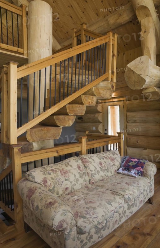 High angle view of twisting staircase with wooden and black wrought iron handrail and brown stained half log steps attached to large supporting central tree trunk post in living room with flowery motifs upholstered sofa inside handcrafted Eastern white pine Scandinavian log cabin home, Quebec, Canada, North America