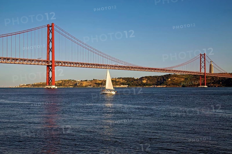 A sailboat passes under a large orange suspension bridge on calm blue waters during evening light, Lissabon, 25 de Abril Bridge