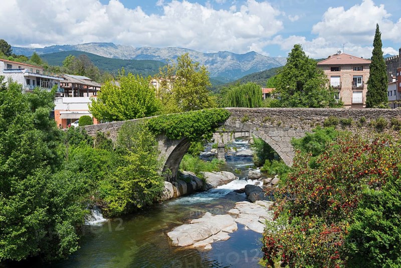 Eine alte Steinbrücke spannt sich über einen Fluss, umgeben von grüner Vegetation und historischen Gebäuden, mit Bergen und blauem Himmel im Hintergrund, Brücke Aquelcabos, Römerbrücke, Puente Romano, Arenas de San Pedro, Fluss Arenal, Ávila, Avila, Kastilien-León, Kastilien-Leon, Spanien