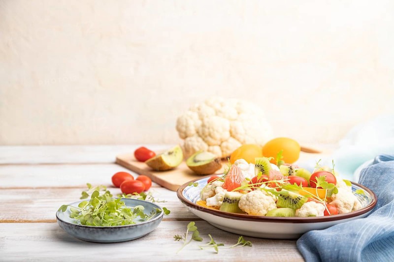 Vegetarian salad of cauliflower cabbage, kiwi, tomatoes, microgreen sprouts on white wooden background and blue linen textile. Side view, copy space, selective focus