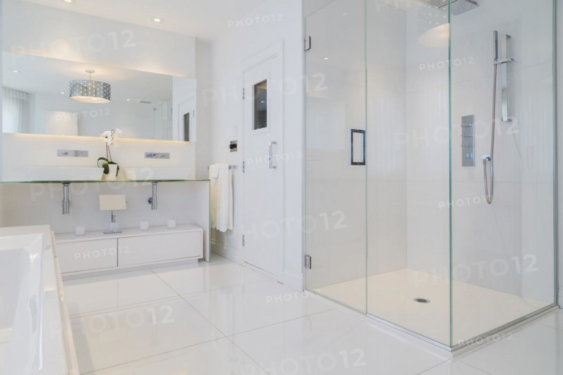 Partial view of white elongated rectangular bathtub and cabinets with large wall mirror, glass shower stall, sauna room door in en suite with ceramic tile flooring on upstairs floor inside a luxurious residential home, Quebec, Canada, North America