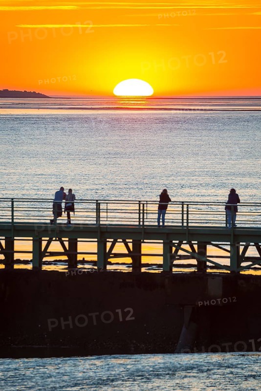 Silhouettes of several people on the pier Freilaufkanal, couple and single persons watch from a jetty the picturesque, golden, breathtakingly beautiful sunset over the sea, island Langlütjen II far away on the horizon, sun half sunk, orange glowing cloudless sky, wide sea in soft colours, calm atmosphere, mouth of the Weser into the North Sea, Outer Weser, Bremerhaven, Land Bremen, Germany, Europe