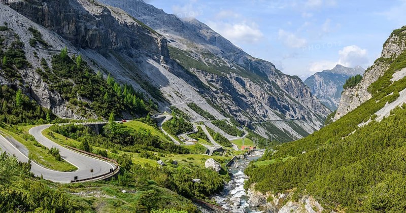 Photo in wide format light panorama of view to south in Braulio valley with foreground right water of waterfall Cascata del Braulio flowing towards in background electric power plant hydroelectric power plant power station Centrale Idroelettrica A2A del Braulio for electricity production production of electricity from hydropower, left pass road south ramp approach to Umbrail Pass and Stelvio Pass Stelvio, Bormio, Lombardy, Italy, Europe