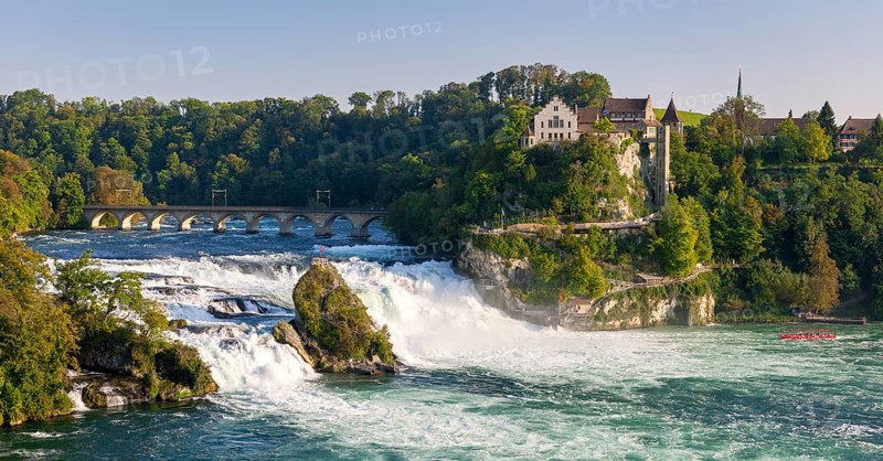 Waterfall flowing next to a historic bridge with lush vegetation and buildings in the background, Schloss Laufen, Rhine Falls, Canton Schaffhausen, Switzerland, Europe