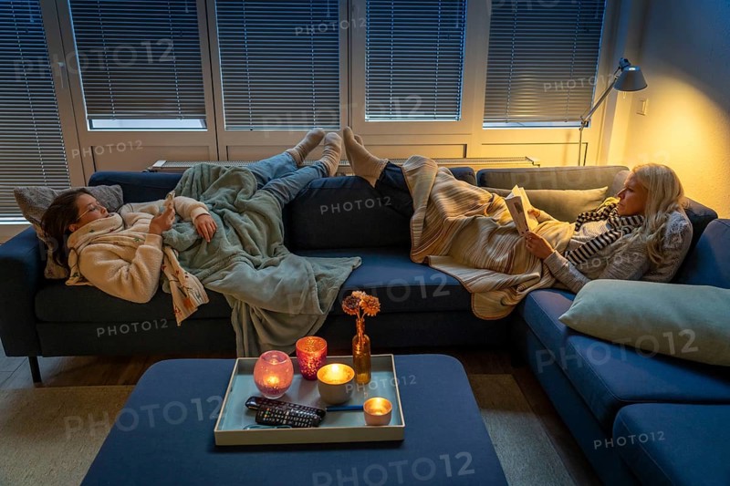 Symbolic image energy saving, cold winter, energy crisis, cold flat, mother and daughter lying on the sofa, warmly dressed, candlelight, heating on to a minimum, warming their feet on the radiator