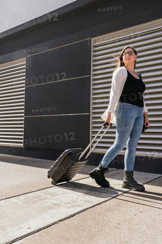 Young business woman pulling rolling suitcase while walking outside airport terminal, preparing to embark on a business trip, representing travel, business, and mobility