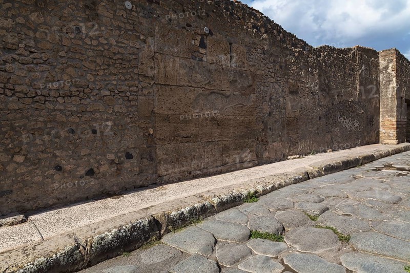 Long narrow stone sidewalk and street made of large grey smooth and worn lava stones plus remnant of old stone and terracotta brick wall structure at the ancient ruins of Pompeii, Campania region, Italy, Europe