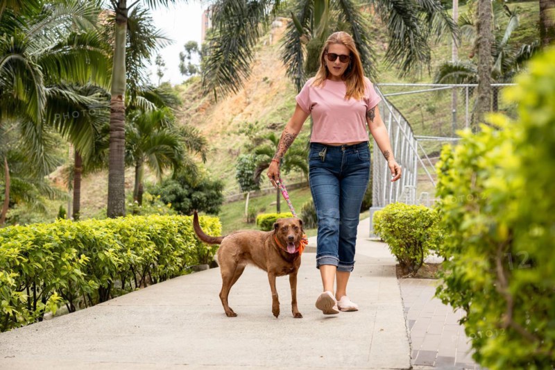 Woman walking her dog, dressed casually in jeans, a pink t-shirt, and sunglasses, in a private neighborhood full of plants and palm trees