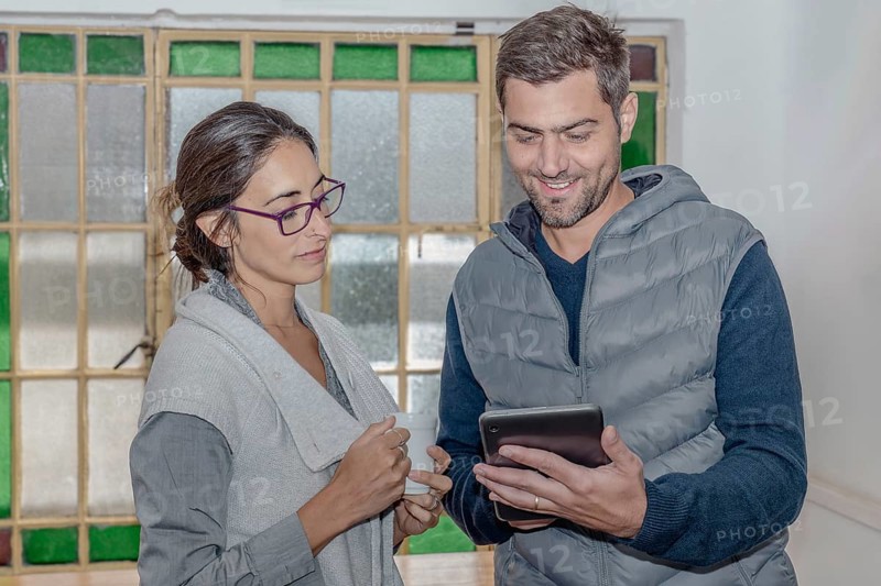 Two colleagues discuss their work looking at a tablet, with friendly expressions and concentrated. The woman holds a cup of coffee. The setting is a brightly lit room with large windows in the background